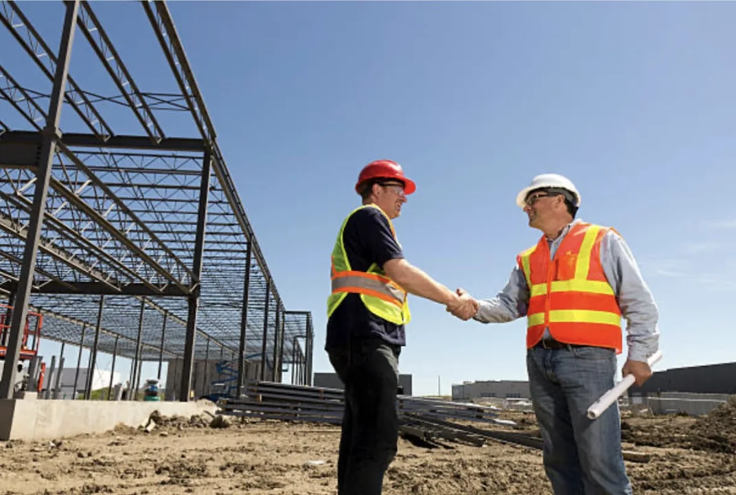 Two construction professionals in hard hats and hi-vis vests shaking hands on site beside a steel-framed industrial building under construction.